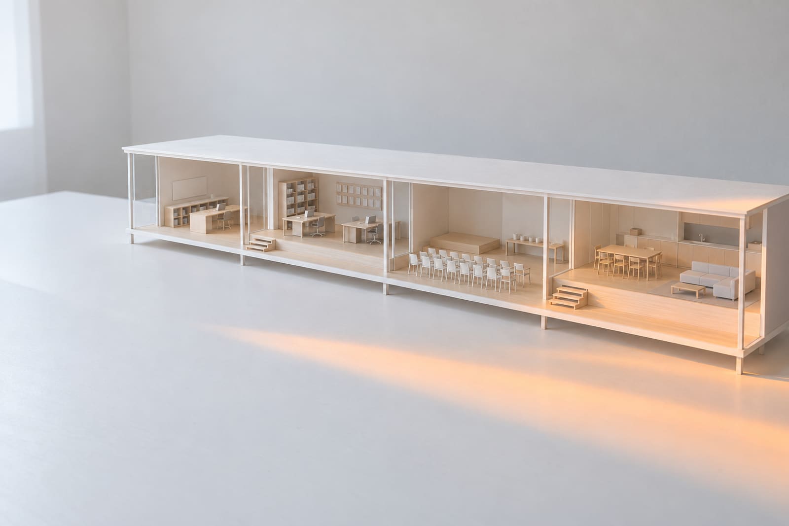 A white card model of a long, low house on a table: a thin white frame, a glass front and four rooms in a row with no doors between them, a studio being set up, an office, a room set for a gathering and a family kitchen.