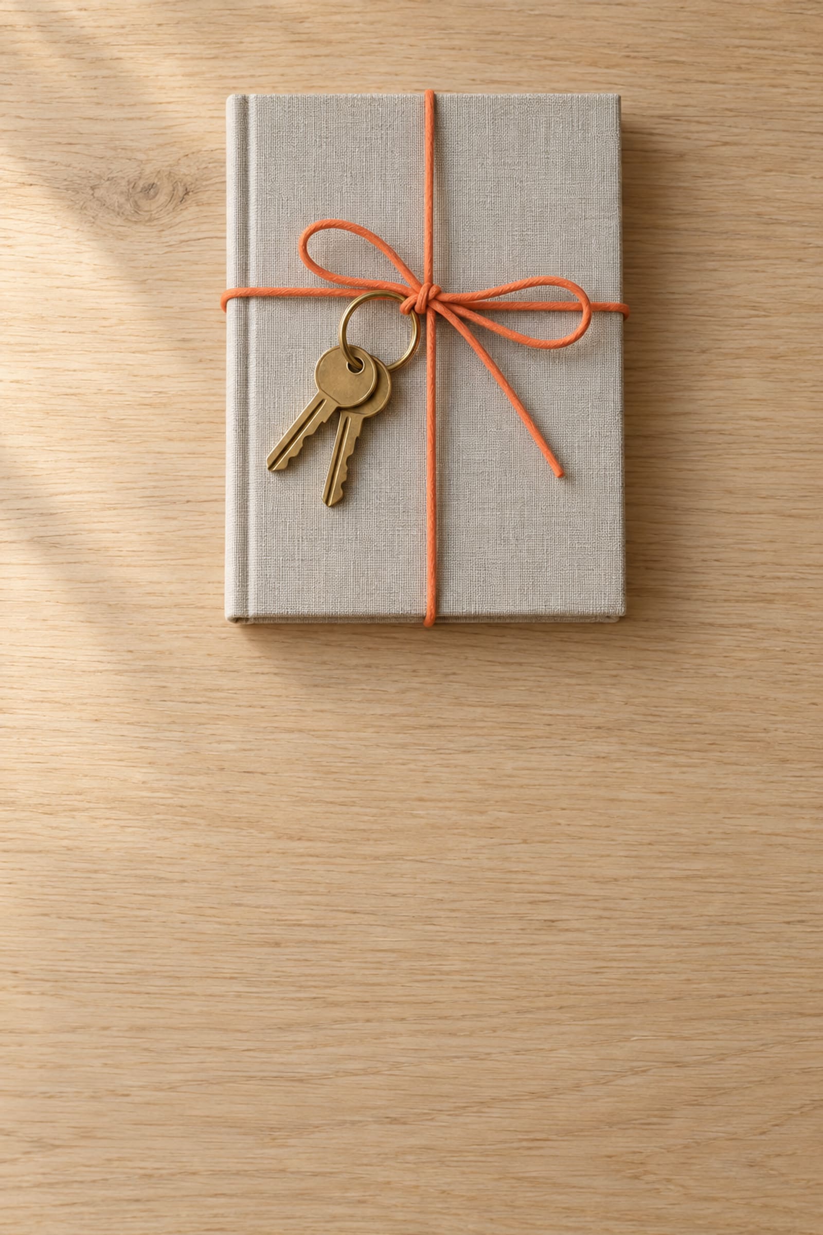 Two brass keys and a grey linen notebook tied together with an orange paper cord, on pale oak.