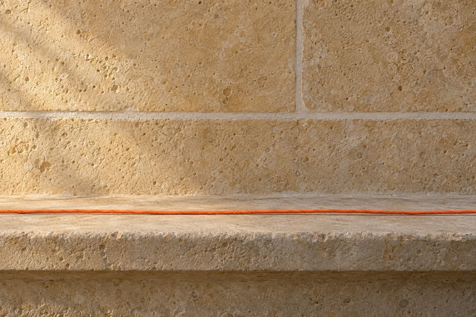 A honey-coloured stone wall in low morning sun, with an orange paper cord lying along its ledge.